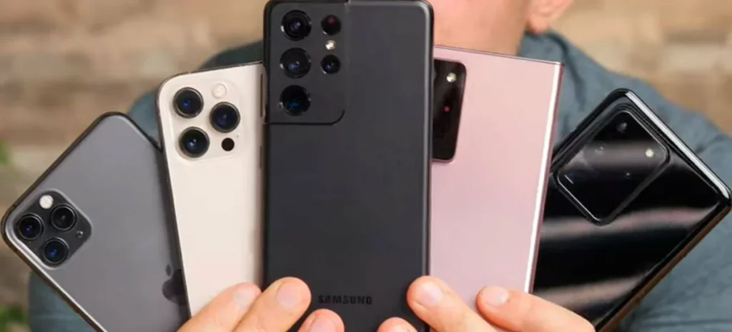 Pessoa segurando cinco smartphones diferentes nas mãos em formato de leque, exibindo os módulos de câmeras traseiras de modelos da Apple e Samsung. A imagem ilustra a diversidade dos melhores celulares de 2026 disponíveis no mercado.